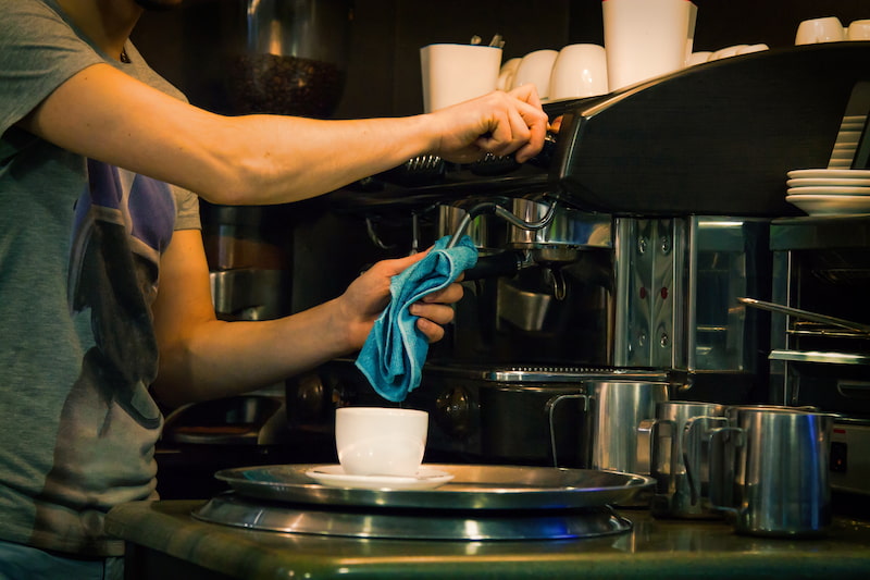 personne préparant un expresso avec une machine à café, tenant un porte filtre et un chiffon bleu, avec une tasse sur un plateau en métal au premier plan.